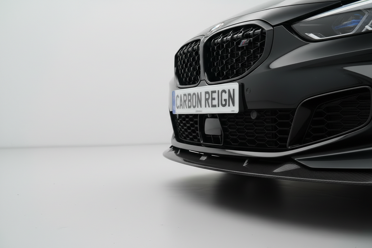 Close-up of the front lower section of a dark gray BMW vehicle, showing the carbon front splitter, honeycomb grille, and a white license plate reading 'CARBON REIGN'.