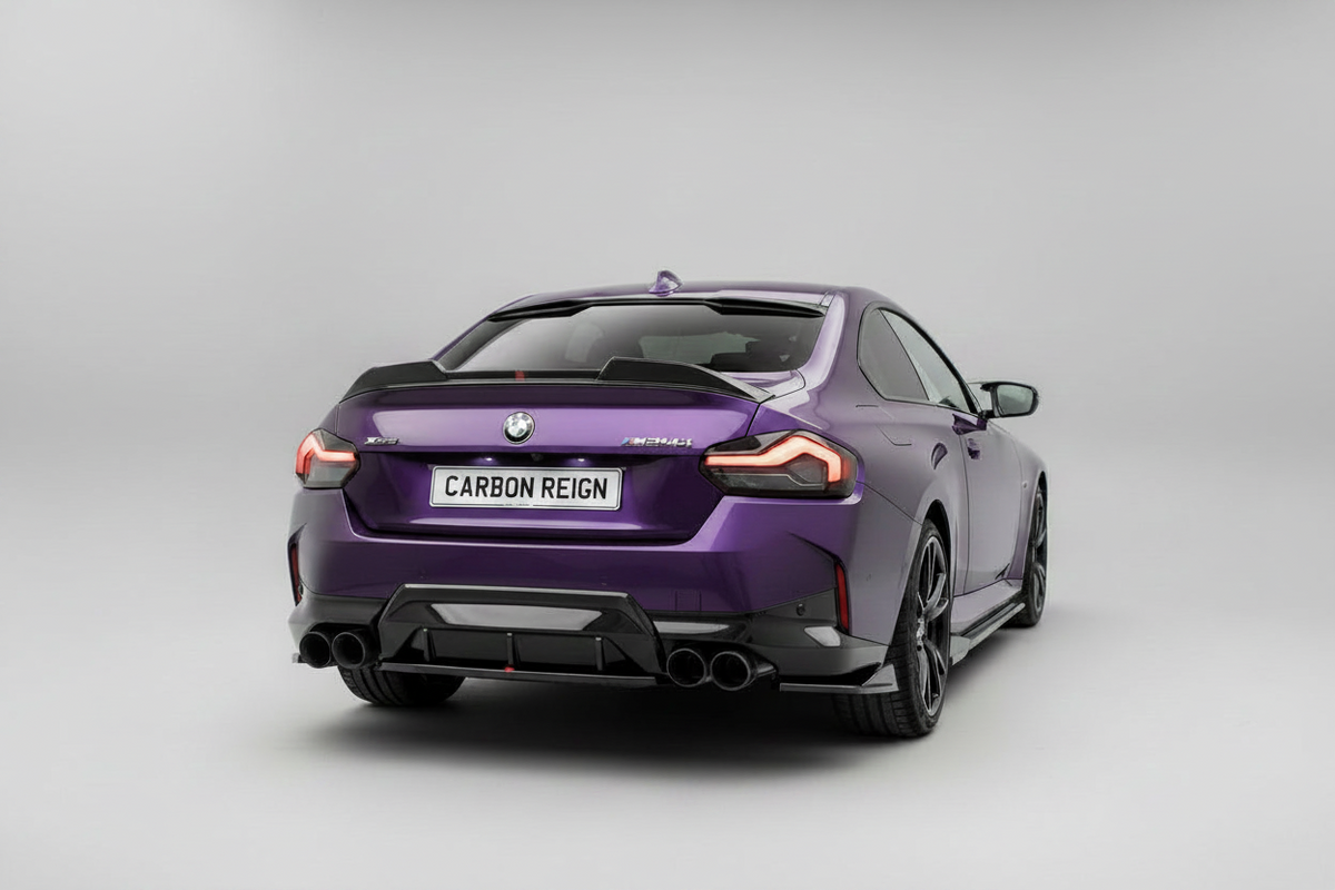 A purple BMW M2 Coupe with a carbon fiber rear diffuser and a license plate reading 'CARBON REIGN', photographed from the rear three-quarter angle on a light gray studio background.