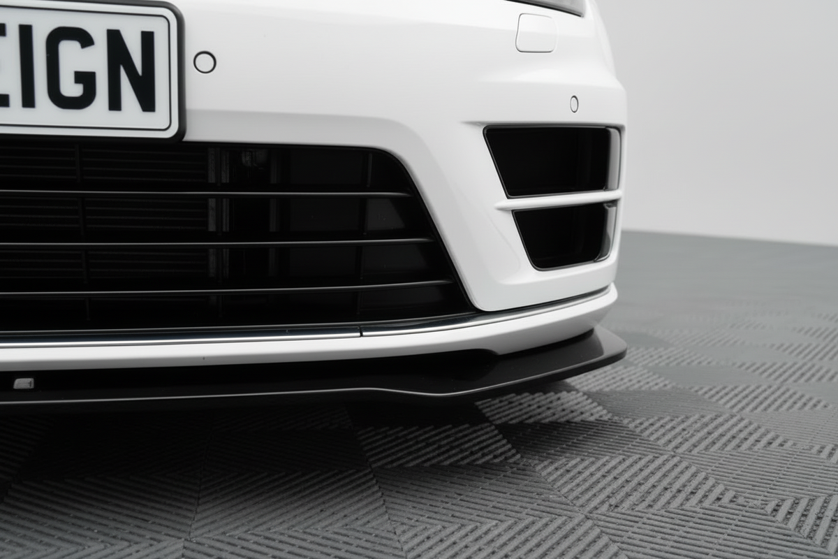 Close-up of the front bumper and lower grille area of a white vehicle, showing a black front splitter, vented air intakes, and part of a dark license plate on a gray textured floor.