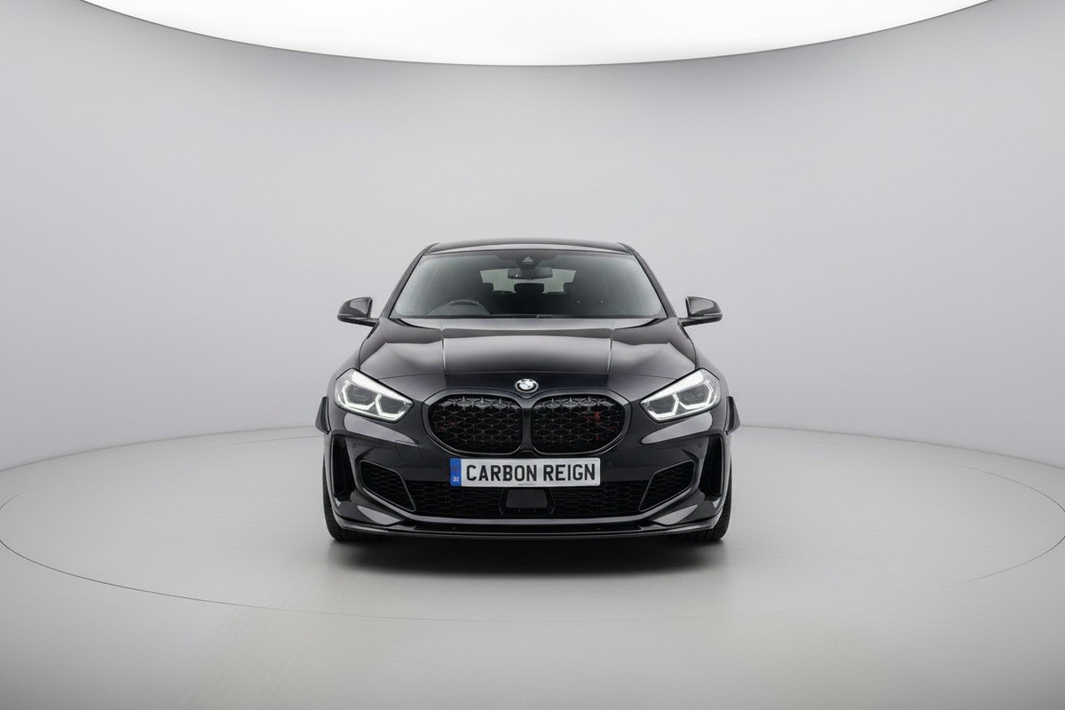 A black BMW 1 Series M2 with a carbon-fiber pattern grille and front splitter, featuring a custom license plate that reads 'CARBON REIGN', photographed from the front in a circular studio setting.